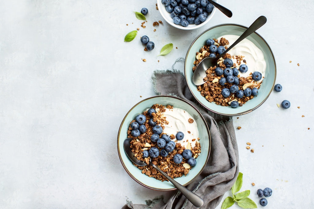 Due ciotole di yogurt con granola e mirtilli freschi, ciascuna con un cucchiaio. Sul tavolo è visibile anche una piccola ciotola di mirtilli e foglie sparse.