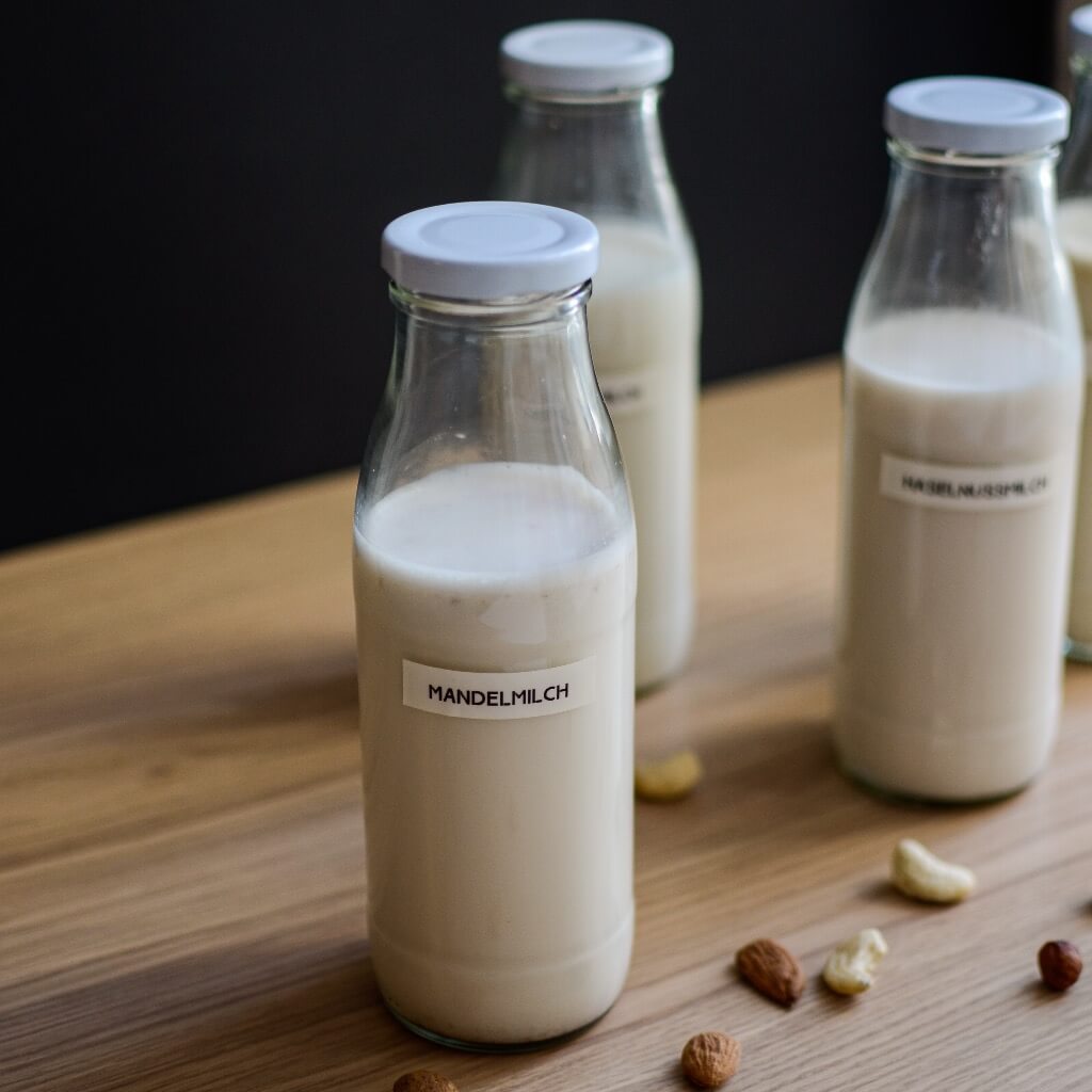 Vier Glasflaschen mit Mandelmilch stehen auf einer Holzoberfläche, umgeben von verstreuten Nüssen. Auf den Flaschen steht „Mandelmilch“.