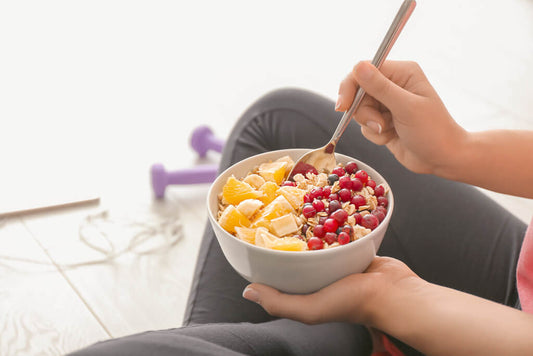 Una persona tiene in mano una ciotola di fiocchi d'avena con fette d'arancia, banana e frutti rossi e si gode questa colazione calda seduta sul pavimento accanto a dei manubri viola, perfetti per chi vuole perdere peso.