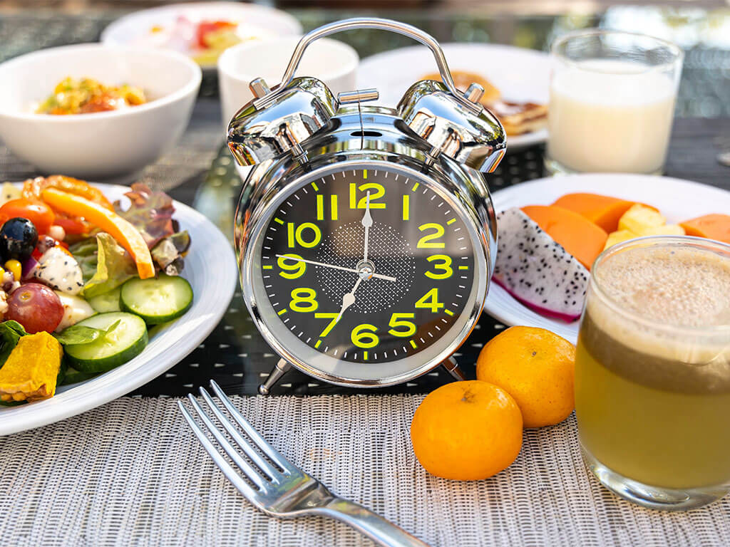 Ein silberner analoger Wecker steht zwischen verschiedenen Frühstücksspeisen, darunter Salat, Obst, Saft und ein Glas Milch auf einem Tisch.