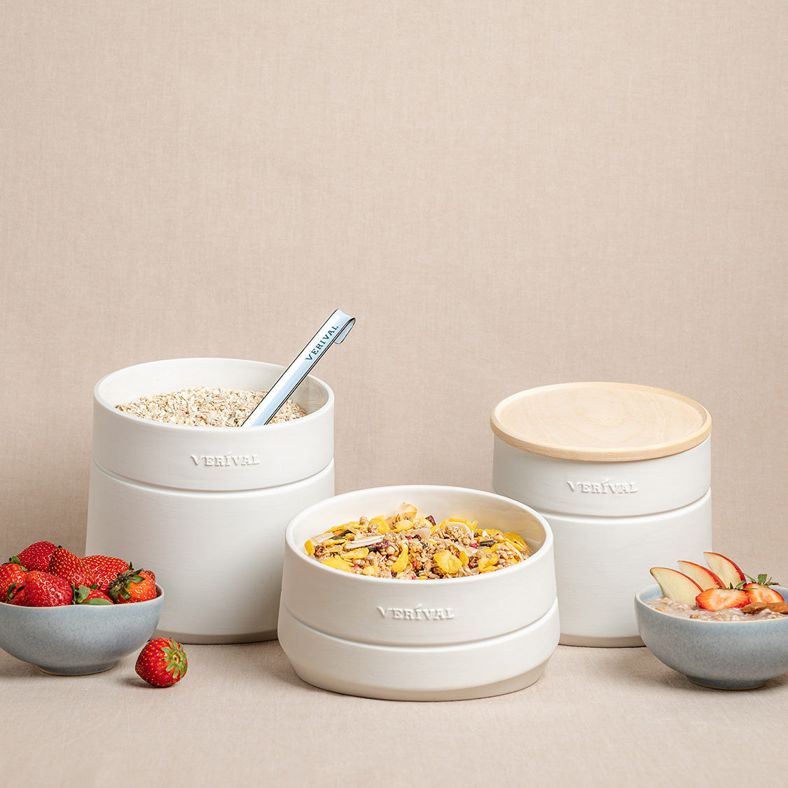 Drei Keramikbehälter, einer mit Müsli und Löffel, einer mit Müsli und Obst und einer mit Holzdeckel, sind mit Schalen mit Erdbeeren und geschnittenem Obst auf einem beigen Hintergrund angeordnet.
