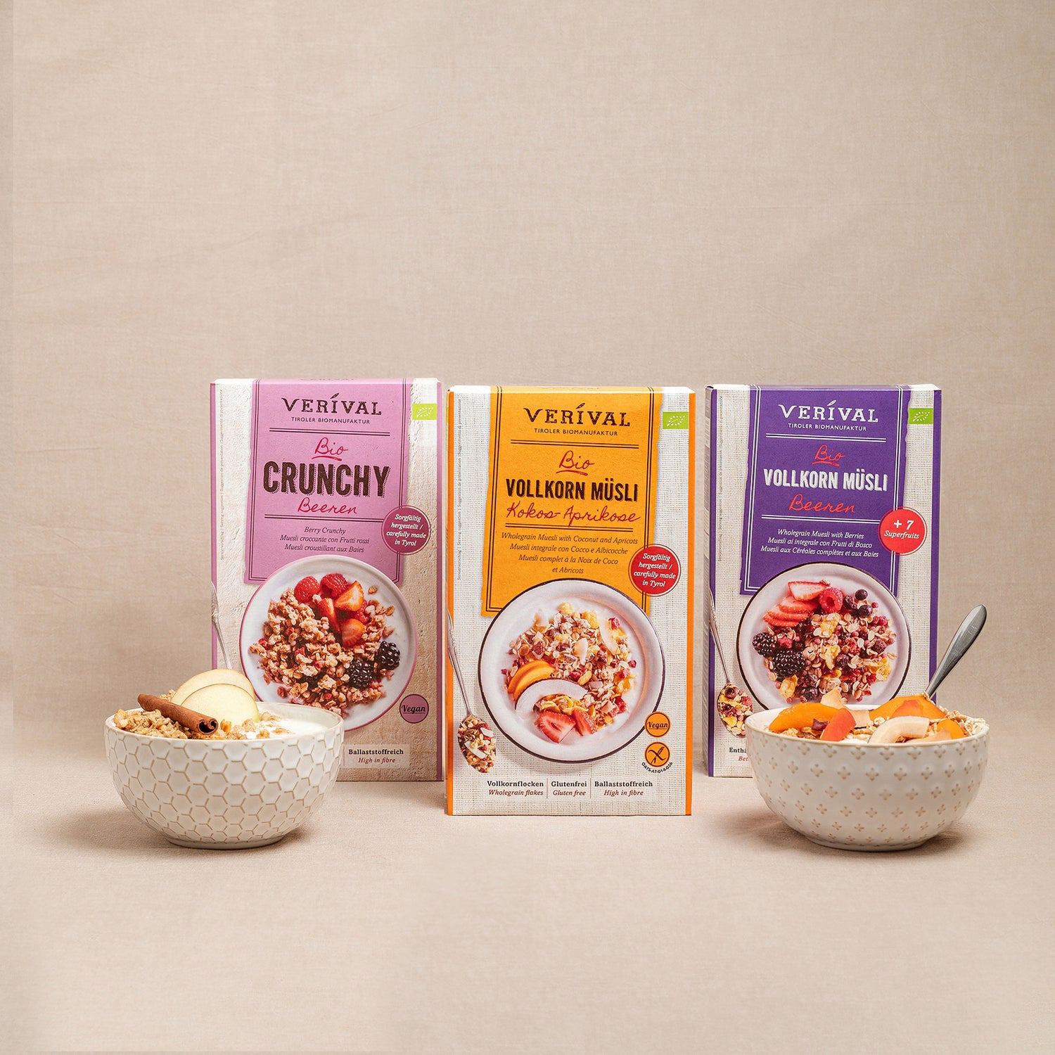 Drei Schachteln mit Verival Müsli und Cerealien stehen hinter zwei Schalen mit Müsli, Obst und Nüssen auf einem beigen Hintergrund.