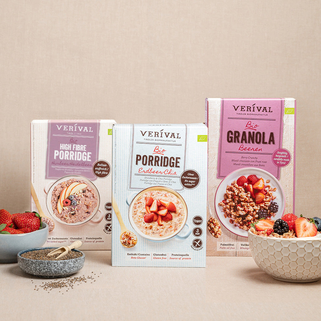 On a beige surface are three Verival cereal boxes with bowls of mixed berries and ready-made muesli, along with a small bowl of chia seeds and a spoon.