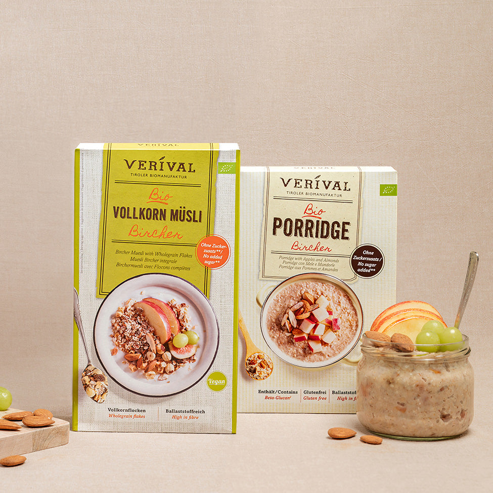 Zwei Schachteln Verival-Müsli, Vollkorn Müsli Bircher und Porridge Bircher, stehen neben einer Dose Müsli mit Früchten und Nüssen auf einer beigen Fläche.