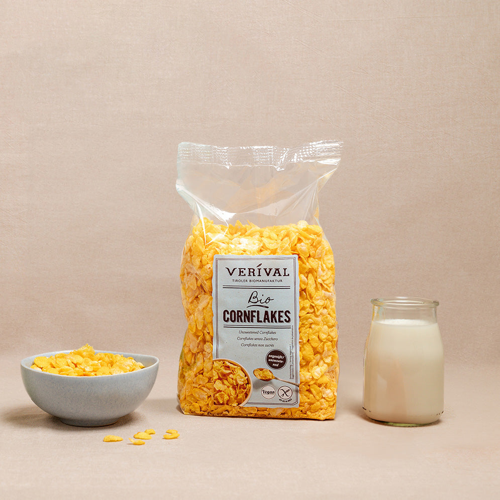 Eine Tüte Verival Bio-Cornflakes, eine kleine Schüssel mit Cornflakes und ein Glas Milch auf einer beigen Fläche mit neutralem Hintergrund.