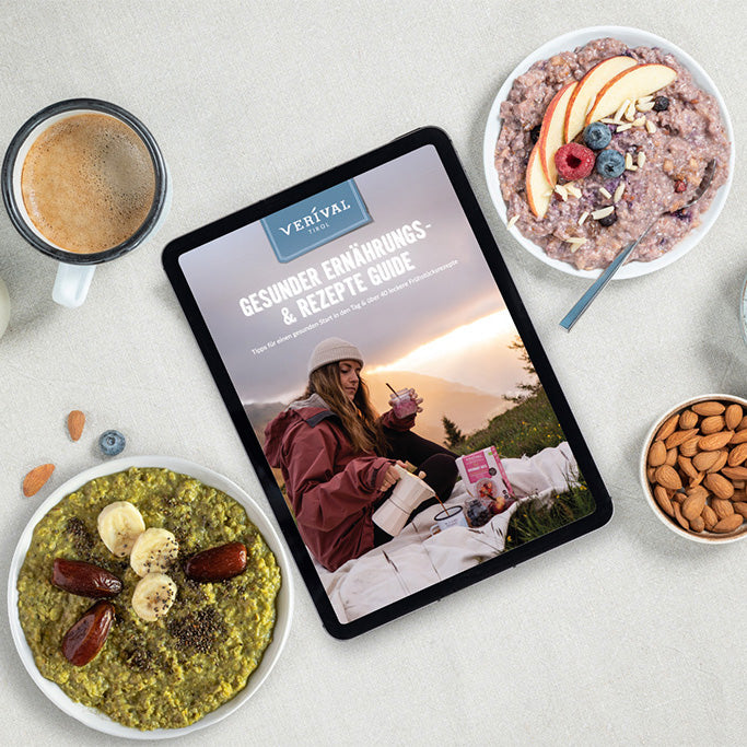 Blick von oben auf einen Tisch mit zwei Schüsseln Brei, einem Kaffee, Mandeln, ein paar verstreuten Beeren und einem Tablet mit einem Ratgeber für gesunde Ernährung und Rezepte.