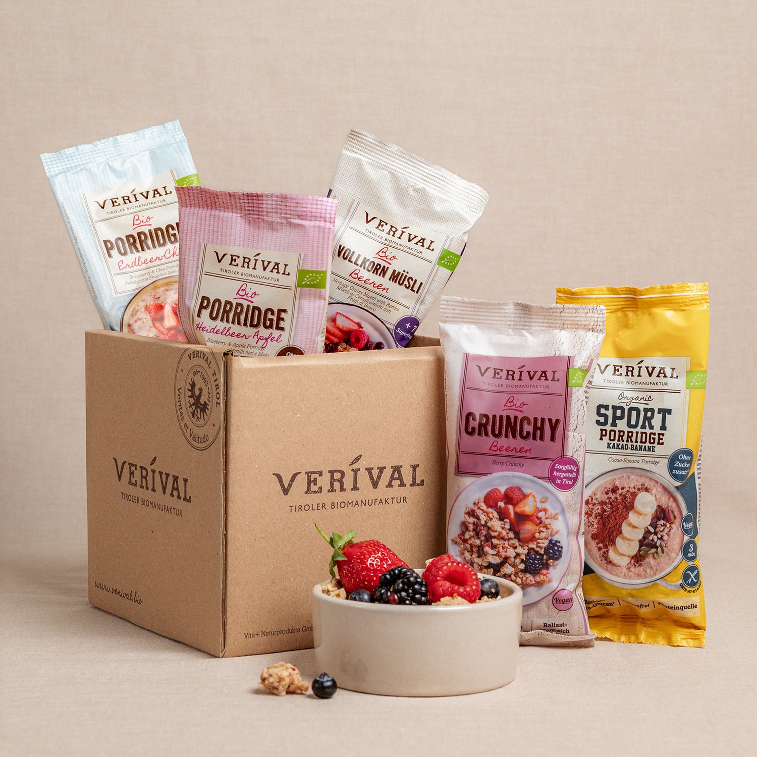Eine Schachtel mit der Aufschrift Verival mit fünf verschiedenen Verival Müsli- und Porridge-Packungen steht neben einer Schüssel mit gemischten Beeren und Müsli auf einem neutralen Hintergrund.