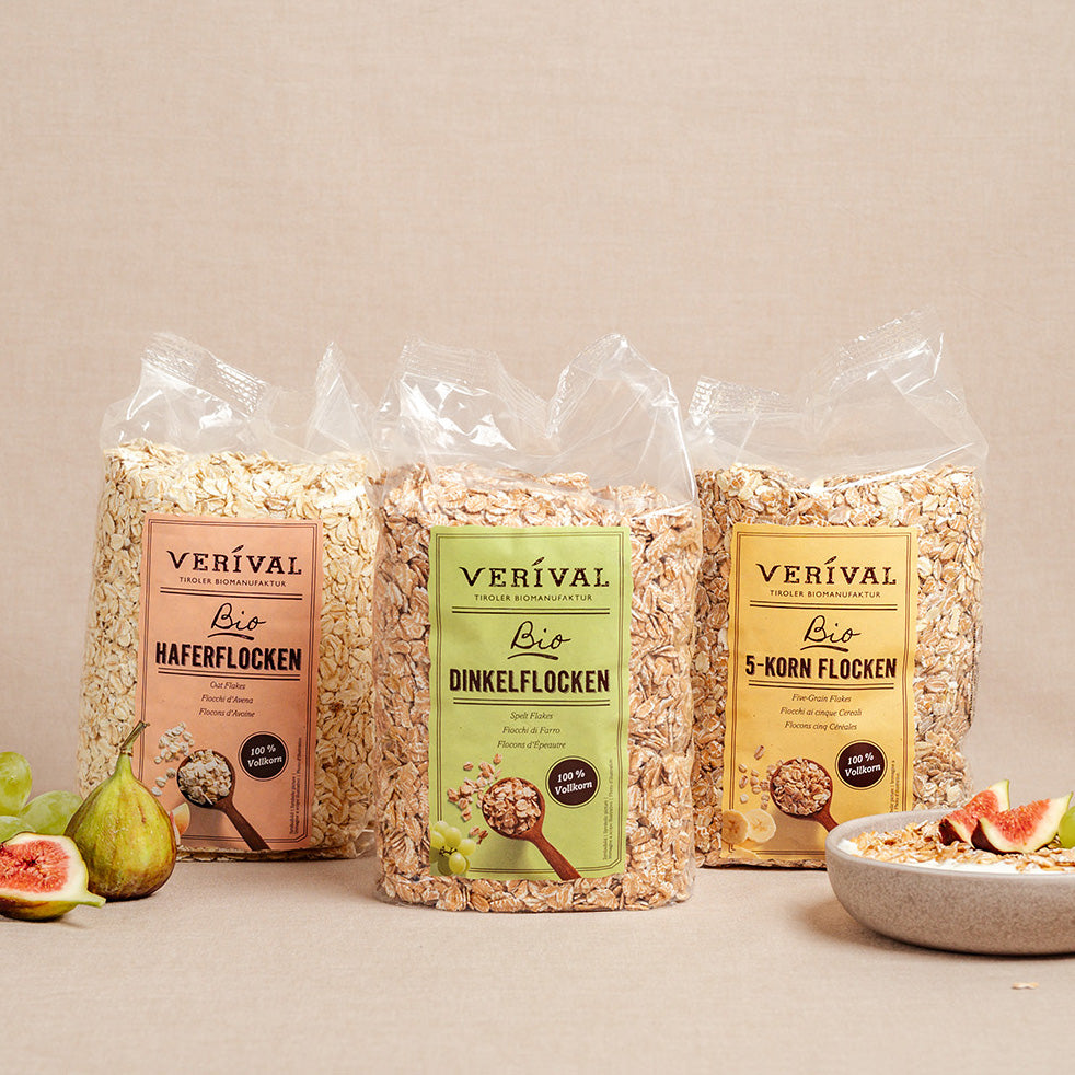 Drei Plastiktüten mit Verival-Getreideflocken - Hafer, Dinkel und 5-Korn - stehen aufrecht auf einer beigen Fläche, daneben stehen Feigen und eine Schale mit Müsli.