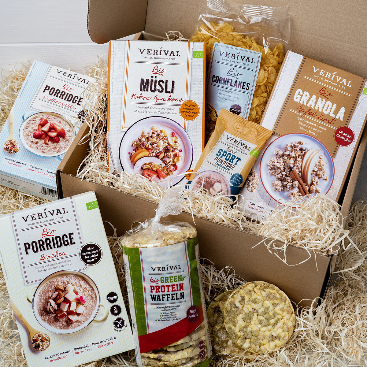 Verschiedene Verival Frühstücksprodukte, darunter Porridge, Müsli, Granola, Cornflakes und grüne Proteinwaffeln, angerichtet in einer Box mit strohähnlichem Verpackungsmaterial.