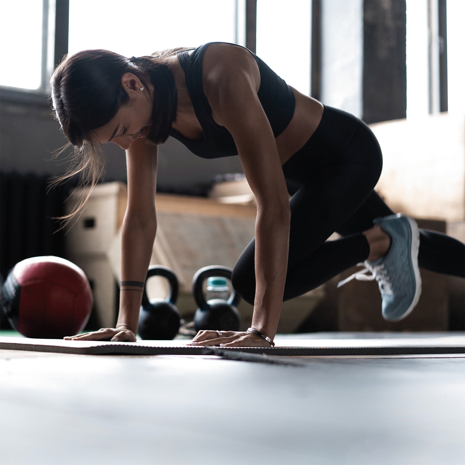 Frau in Sportkleidung bei einer Bergsteigerübung auf einer Matte in einem Fitnessstudio, mit Kettlebells und einem Medizinball im Hintergrund.
