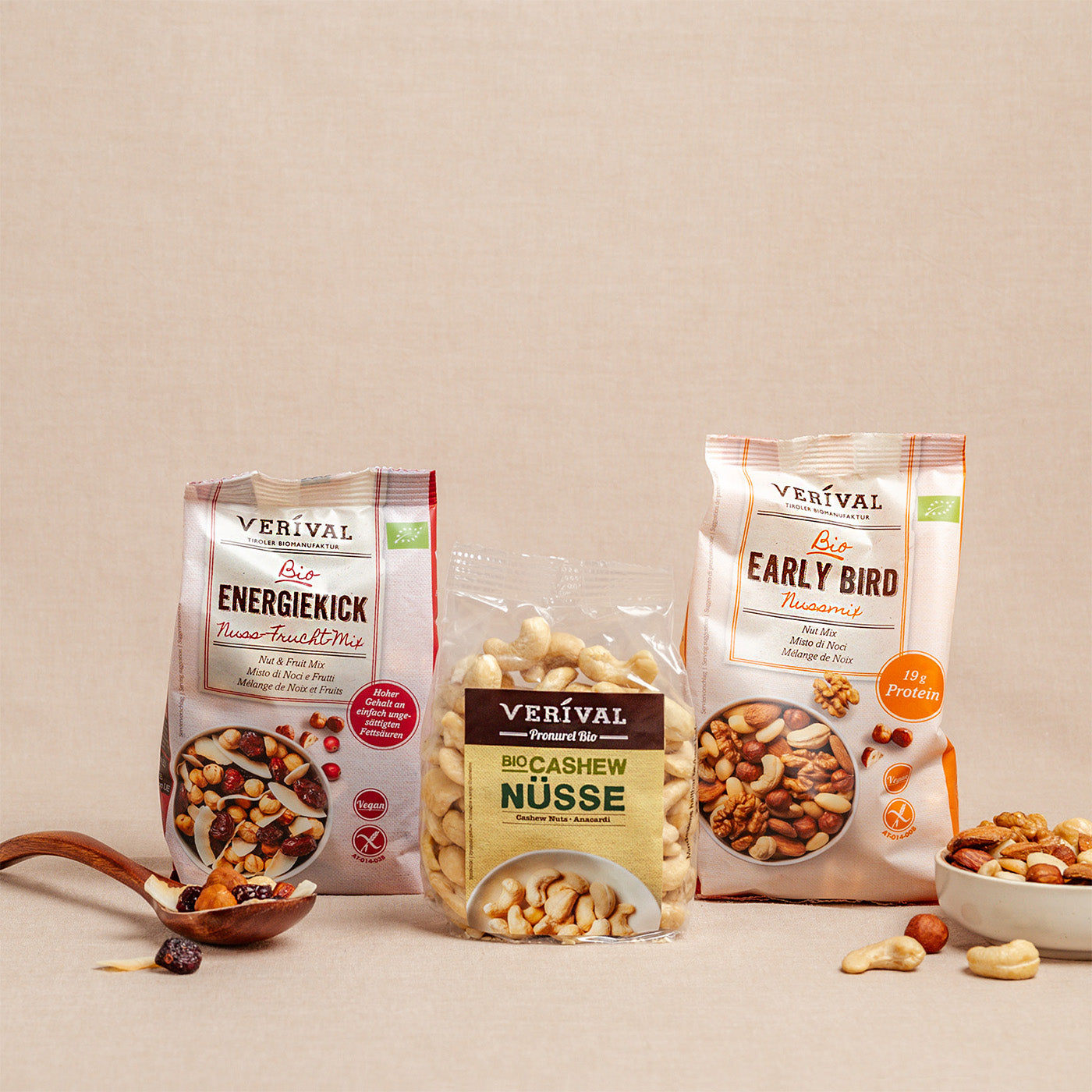 Auf einer beigen Fläche sind drei Verival-Snackpakete ausgestellt: zwei Nuss-Frucht-Mischungen und eine Tüte Cashewnüsse, mit einem Holzlöffel und verstreuten Nüssen im Vordergrund.