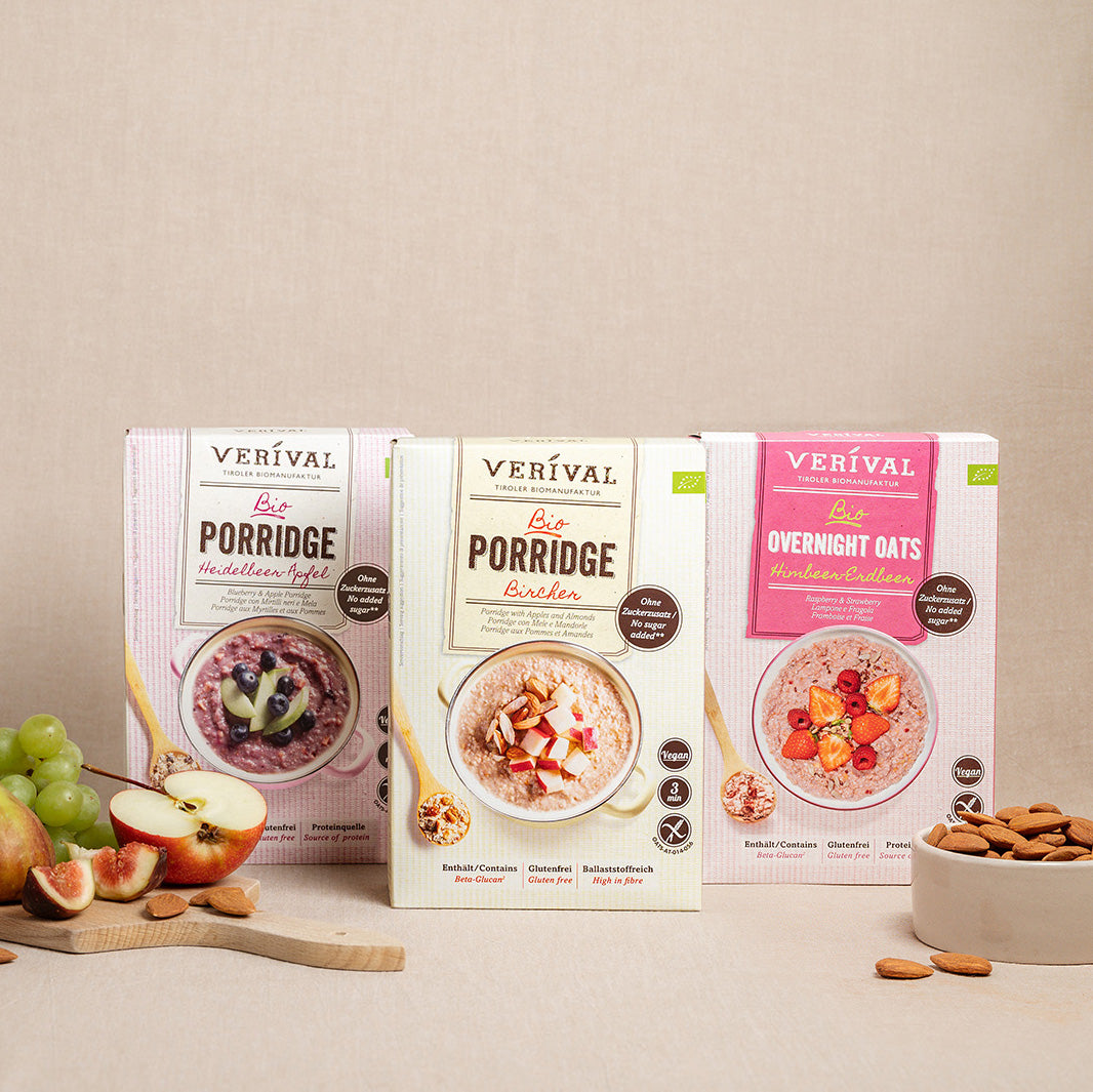 Drei Verival-Frühstücksdosen - Heidelbeer Apfel Porridge, Bircher Porridge und Himbeer-Cranberry Overnight Oats - werden mit Obst und Nüssen auf einer beigen Fläche präsentiert.