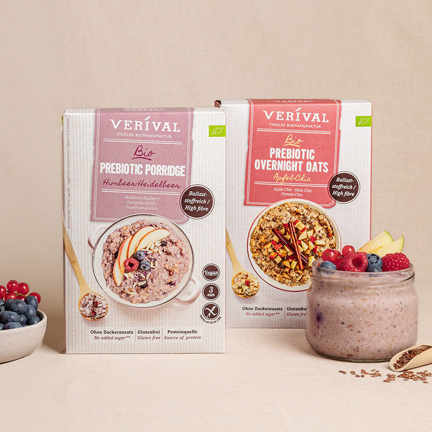 Zwei Schachteln präbiotische Verival-Cerealien und ein Glas zubereitete Haferflocken mit frischen Beeren auf einer neutralen Unterlage, daneben eine kleine Schale mit Blaubeeren.