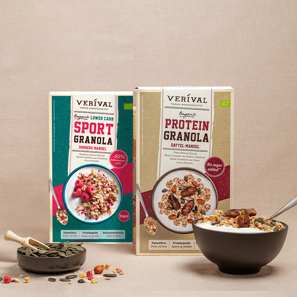 Zwei Schachteln Verival-Granola, eine mit der Aufschrift Sport Granola und die andere mit der Aufschrift Protein Granola, stehen neben einer Schüssel mit Müsli und verstreuten Samen und Nüssen.