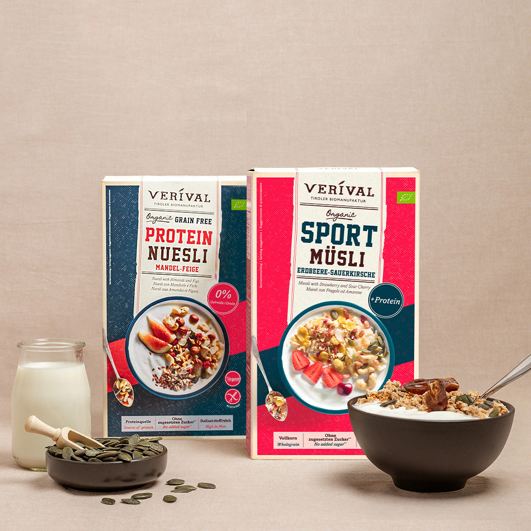 Zwei Schachteln Verival-Müsli, eine Schale Müsli mit Obst, ein Glas Milch, ein Löffel und eine kleine Schale mit Körnern auf einer beigen Fläche.