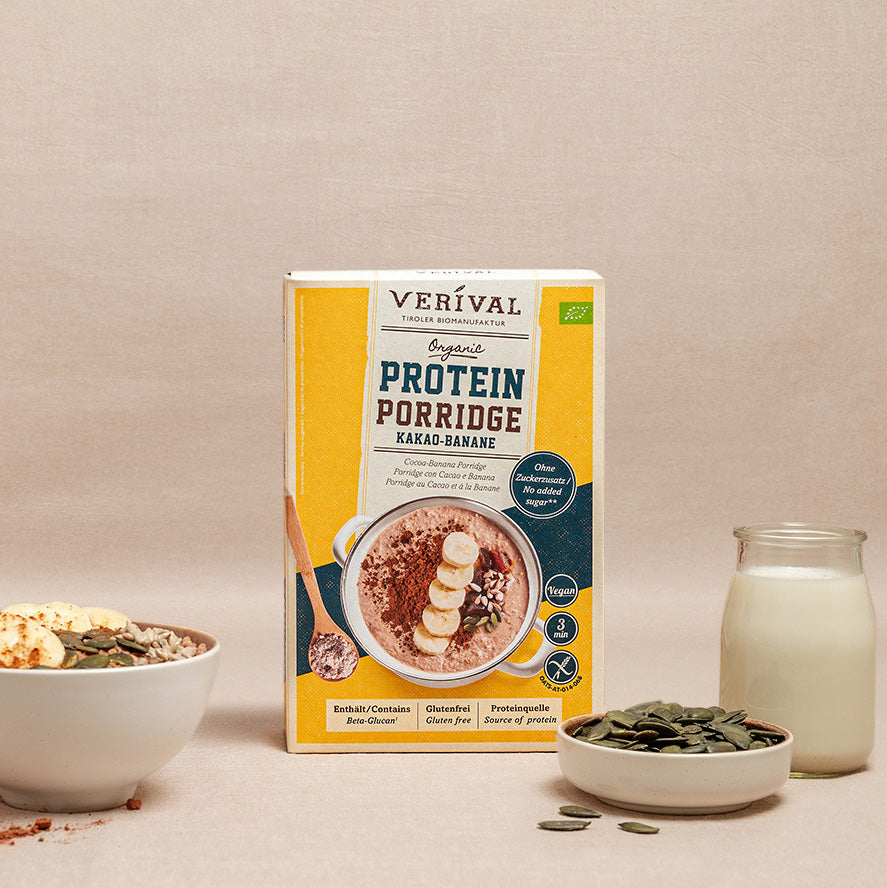 Eine Schachtel Verival Bio-Proteinbrei wird mit einer Schüssel Brei mit Bananenscheiben und Kernen, einem Glas Milch und einer kleinen Schale mit Kürbiskernen präsentiert.