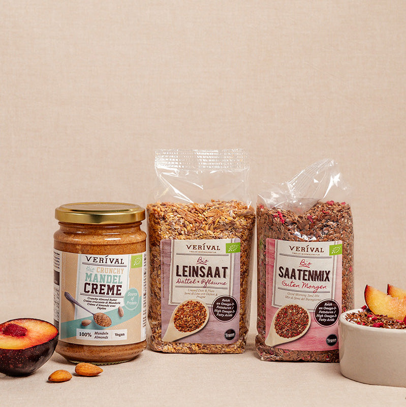 Drei vegane Produkte der Marke Verival - Mandelcremeaufstrich, Leinsamen und Saatenmischung - sind auf einer beigen Fläche ausgestellt, daneben eine Pflaume, Mandeln und eine Schale Müsli.