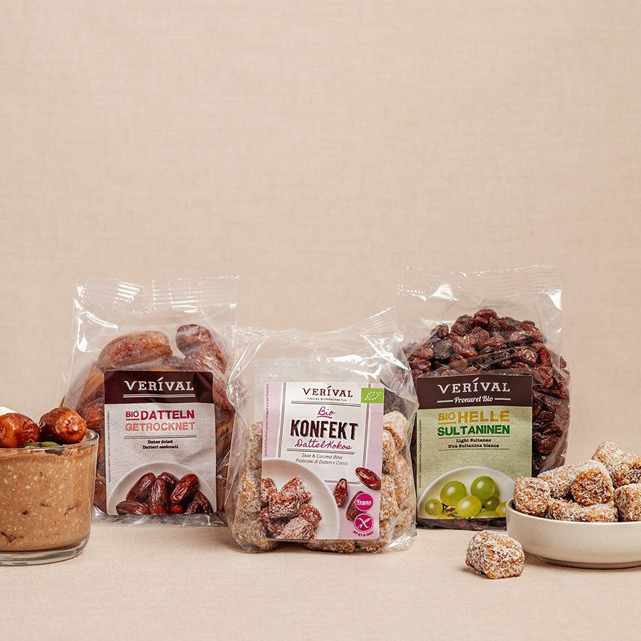 Drei Snack-Packungen der Marke Verival - getrocknete Datteln, Süßwaren-Happen und leichte Rosinen - sind in einer Reihe angeordnet, mit einer Schale mit Brotaufstrich und Energie-Happen im Vordergrund.