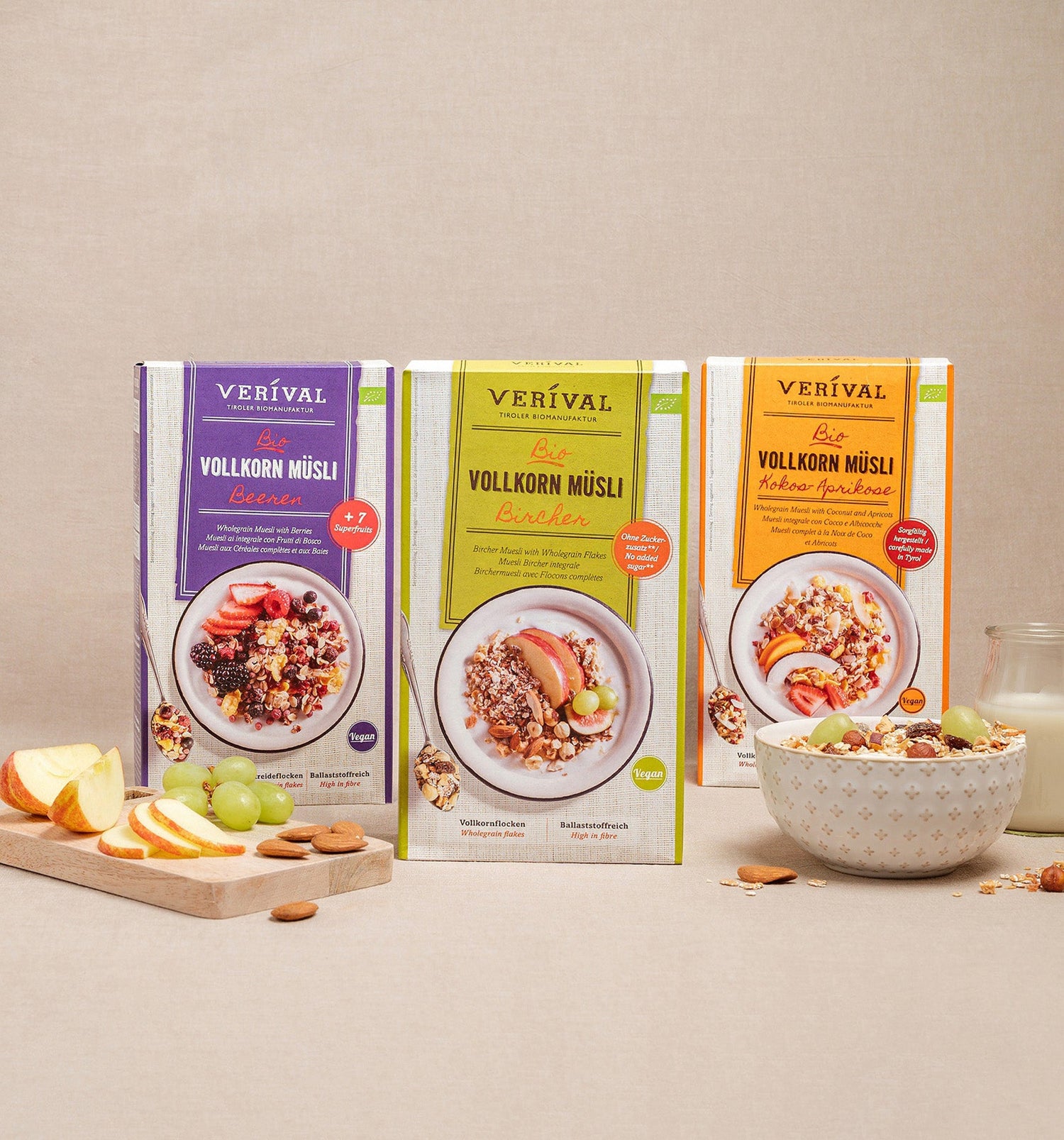 Tre scatole di muesli integrale Verival in vari gusti sono disposte verticalmente su una superficie beige, accanto a una ciotola di muesli, frutta e un bicchiere di latte.