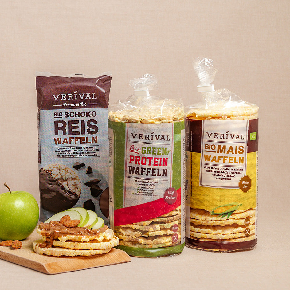 Drei Packungen Reis- und Maiskuchen der Marke Verival stehen aufrecht neben einem grünen Apfel, geschnittenen Apfelstückchen und einigen Mandeln auf einem Holzbrett.