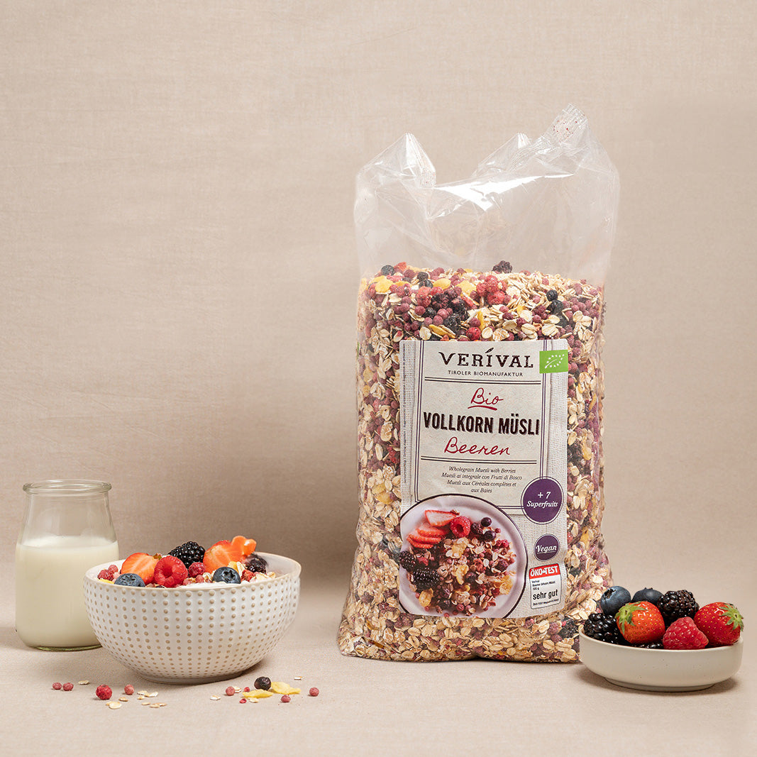 Eine Tüte Verival Bio Vollkorn Müsli Beeren steht neben einer Schale Müsli mit Beeren und einem Glas Milch, in einer kleinen Schale daneben befinden sich weitere Beeren.