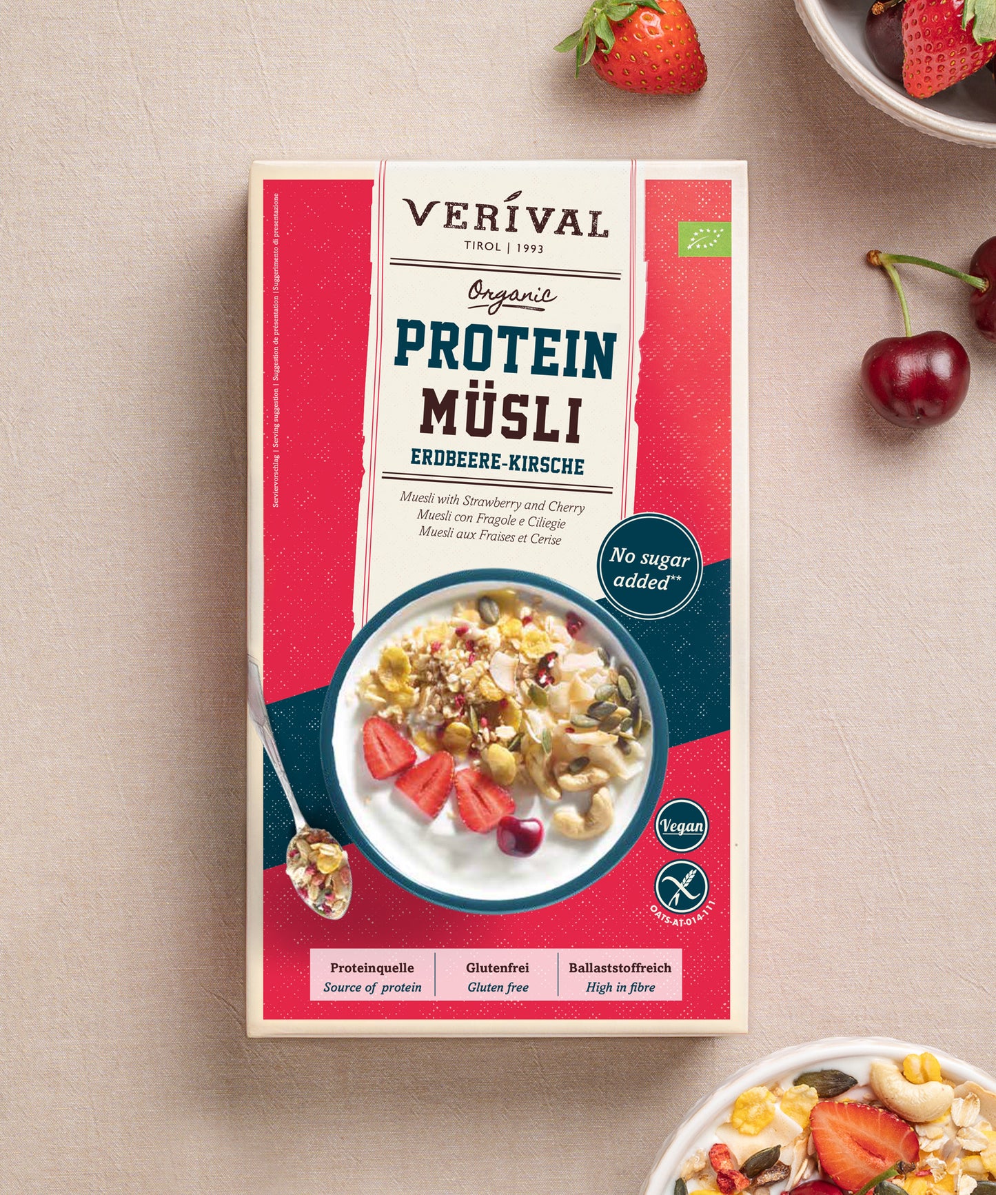 Eine Schachtel des Bio Protein Müsli Erdbeere-Kirsche der Verival Bio GmbH wird auf einer hellen Fläche mit frischen Erdbeeren und Kirschen präsentiert. Dieses Müsli ist eine pflanzliche Proteinquelle und enthält keinen Zuckerzusatz.