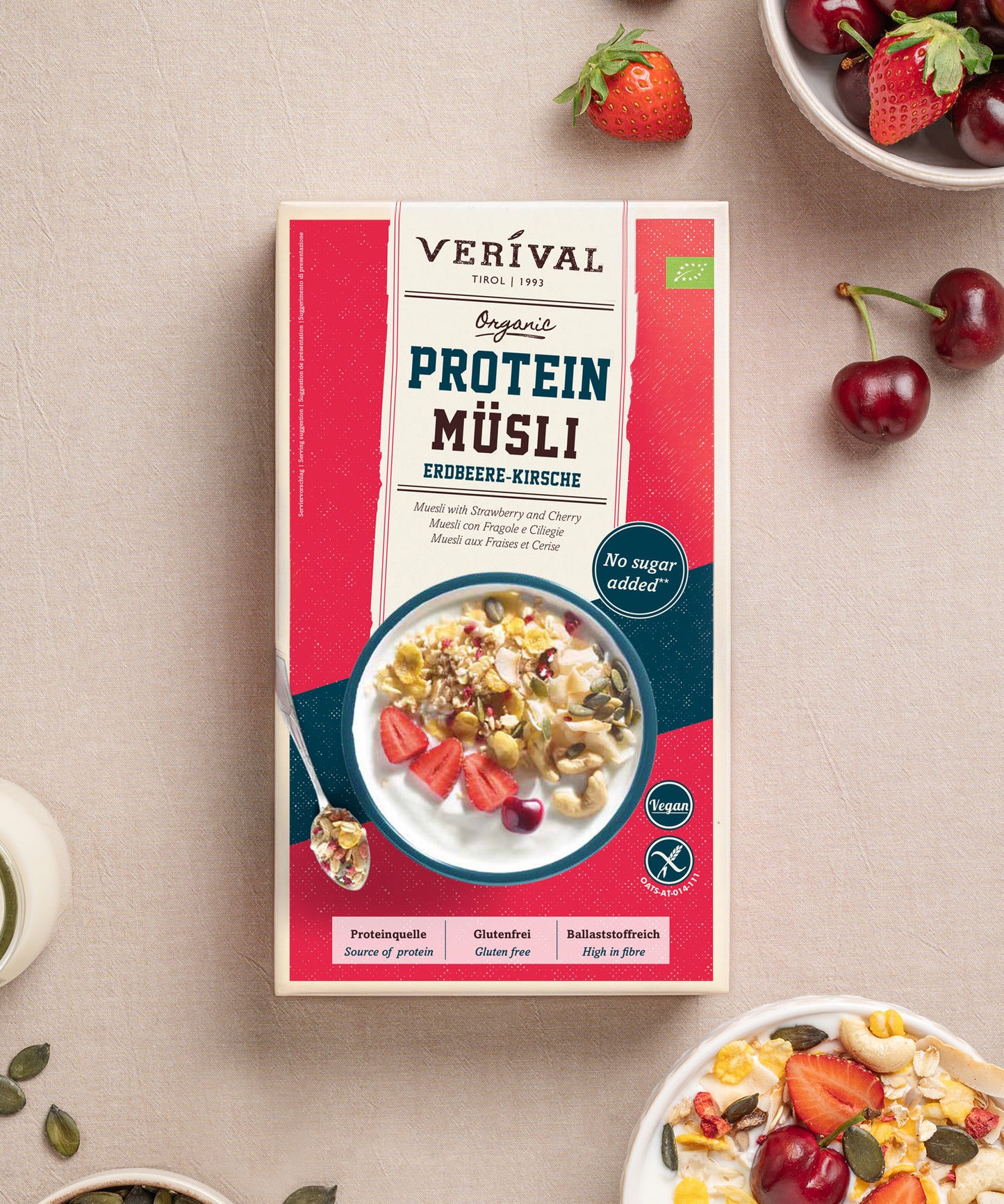 Eine Schachtel Verival Bio GmbH Bio Protein Müsli Erdbeere-Kirsche steht auf einer beigen Fläche, umgeben von Schalen mit Müsli und frischem Obst - ohne Zuckerzusatz und reich an pflanzlichem Protein.
