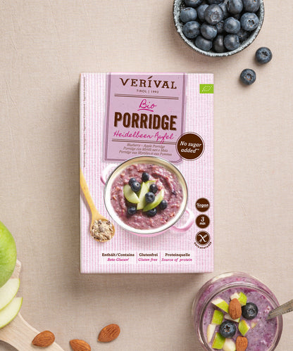 Eine Schachtel Verival Bio GmbH Bio Heidelbeer-Apfel Porridge steht auf einem Tisch mit einer Schüssel veganem, ballaststoffreichem Porridge, umgeben von frischen Heidelbeeren, Apfelscheiben und Mandeln.