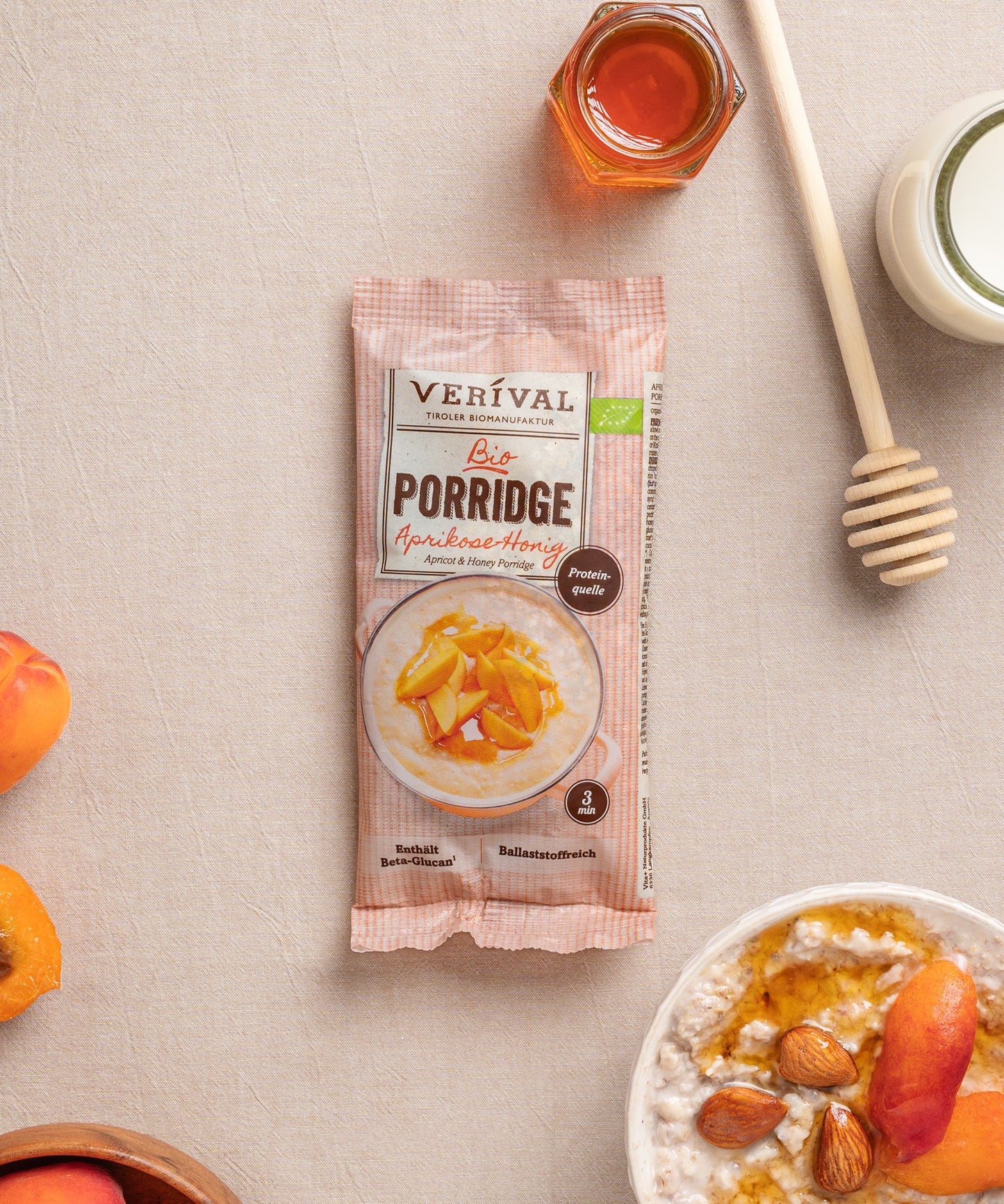 Eine einzelportionspackung Aprikosen-Honig Porridge von Verival Bio, umgeben von Aprikosen, Mandeln, einem Honiglöffel und einem Glas Honig. Laktosefrei und ballaststoffreich – für einen gesunden Start in den Tag.