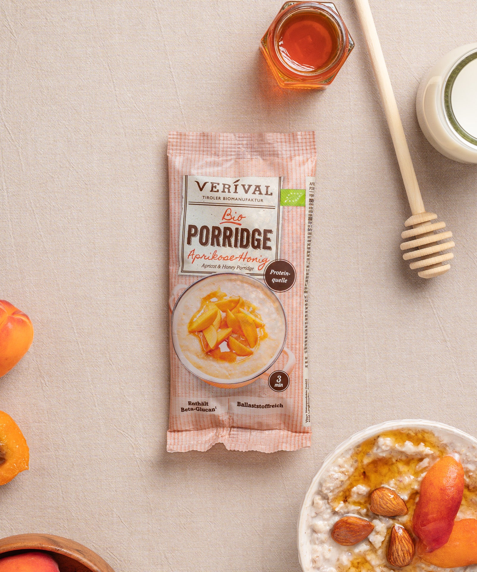 Eine einzelportionspackung Aprikosen-Honig Porridge von Verival Bio, umgeben von Aprikosen, Mandeln, einem Honiglöffel und einem Glas Honig. Laktosefrei und ballaststoffreich – für einen gesunden Start in den Tag.