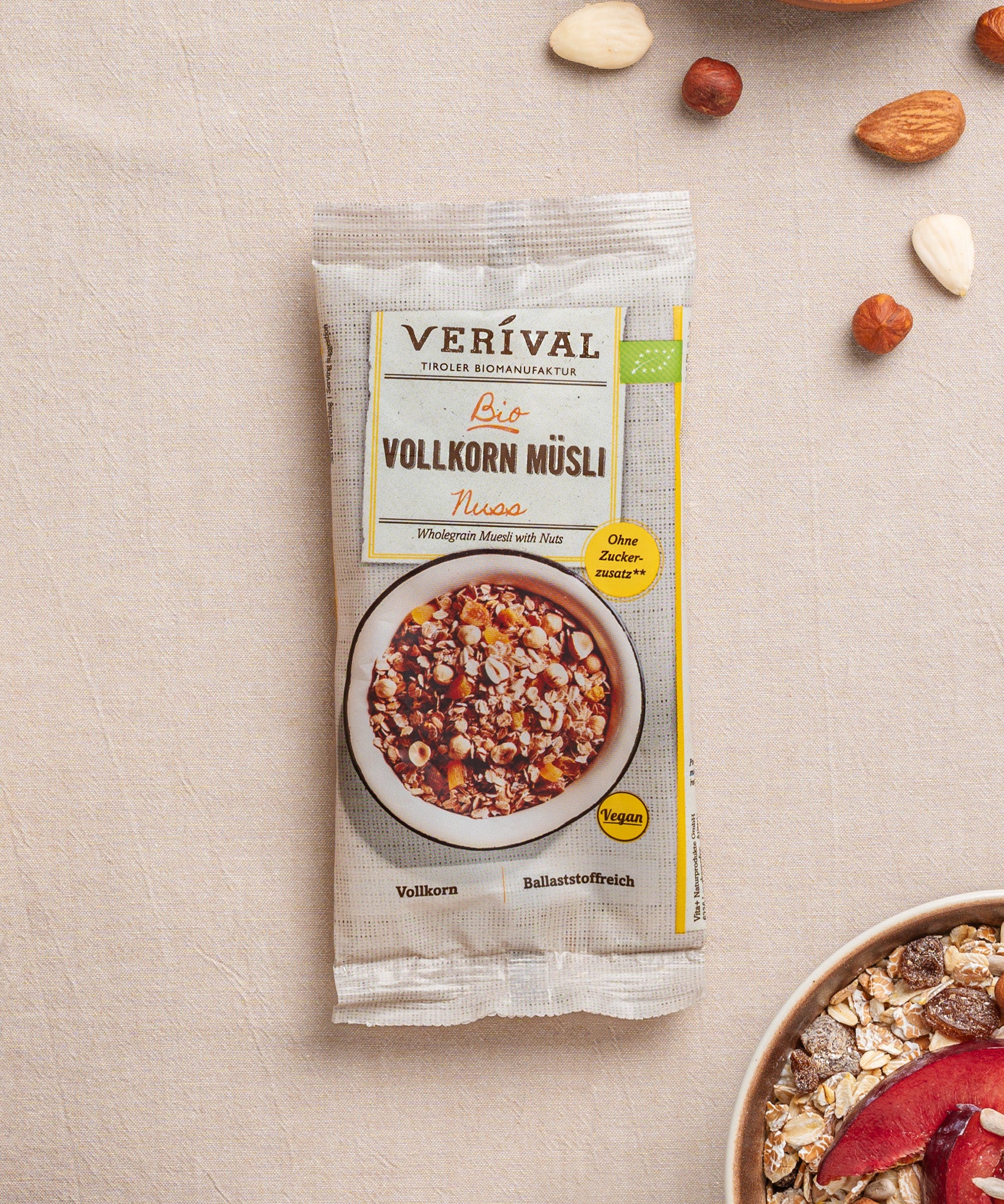 Eine Packung Nuss-Vollkorn-Müsli der Verival Bio GmbH liegt auf einer strukturierten Oberfläche und unterstreicht so seinen veganen Reiz. Die Schachtel enthält eine Schale ballaststoffreiches Müsli mit Nüssen, dazu verstreute Nüsse und einen Teil einer weiteren Schale.