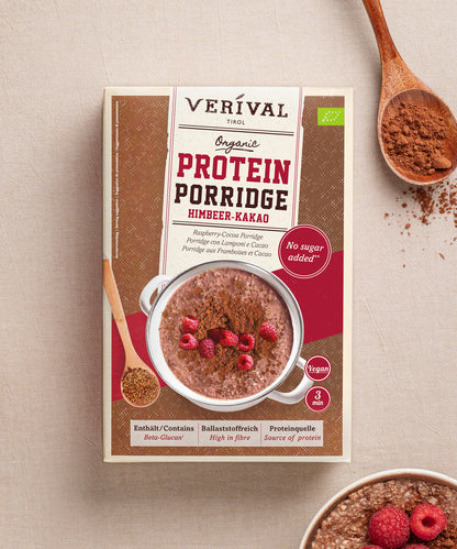 Eine Box Protein Porridge Himbeer-Kakao der Verival Bio GmbH präsentiert sich mit einer Schale veganem Porridge, frischen Himbeeren und Kakaopulver – ein ideales Power-Frühstück für den Start in den Tag.