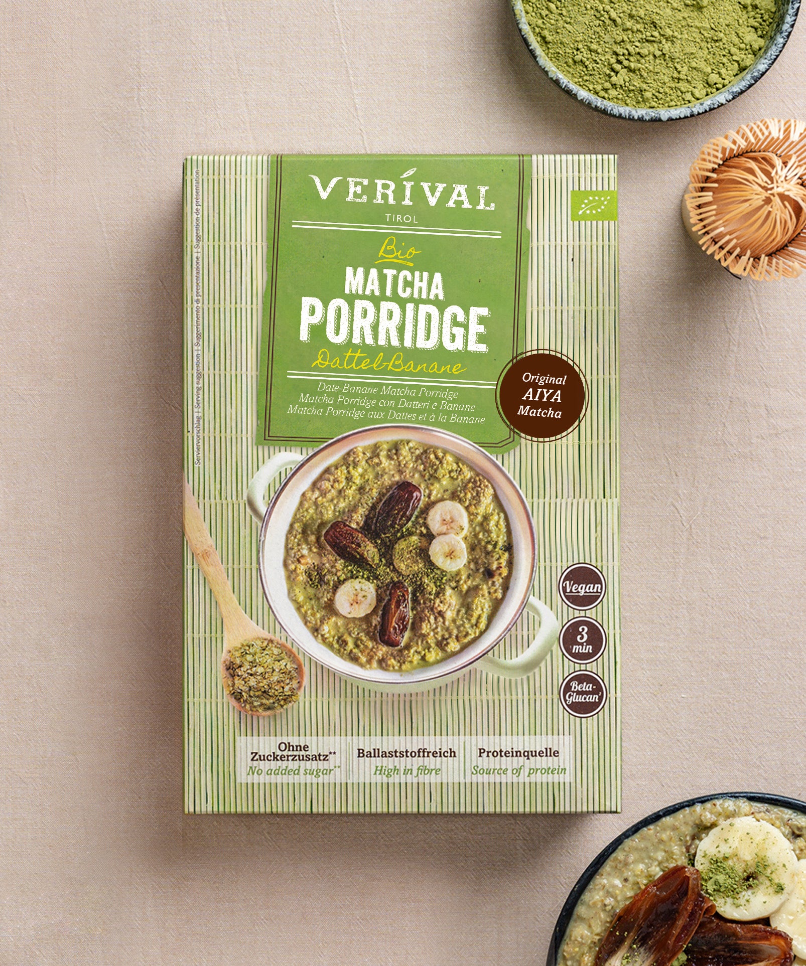 Eine Schachtel Bio Matcha Porridge Dattel-Banane der Verival Bio GmbH steht auf einer beigen Oberfläche, begleitet von Matcha-Pulver, einem Löffel und einer kleinen Schüssel veganem Porridge.