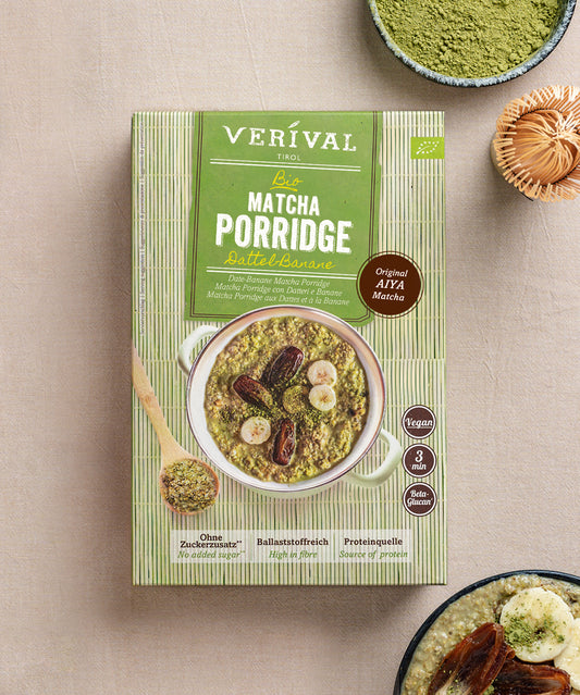 Eine Schachtel Bio Matcha Porridge Dattel-Banane der Verival Bio GmbH steht auf einer beigen Oberfläche, begleitet von Matcha-Pulver, einem Löffel und einer kleinen Schüssel veganem Porridge.
