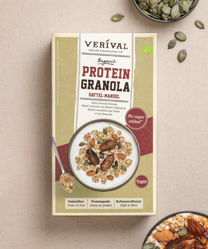 Eine Packung Protein-Granola Dattel-Mandel von Verival Bio, reich an pflanzlichen Proteinen, steht auf einer Tischdecke, umgeben von Schüsseln mit dem ballaststoffreichen Granola, umgeben von verstreuten Mandeln und Datteln.