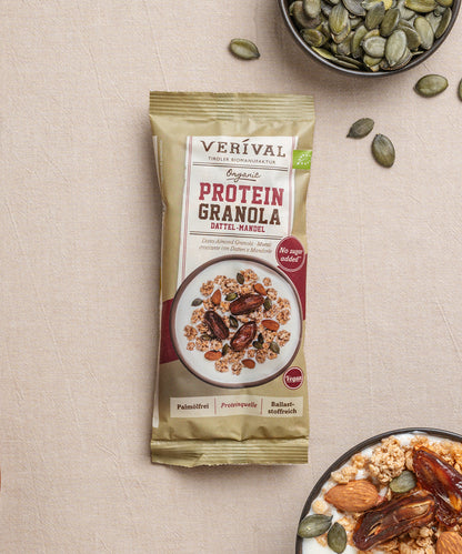 Eine Enzelportionspackung Protein-Granola Dattel-Mandel von Verival Bio steht neben einer Joghurtschale mit Granola, Mandeln, Datteln und Kürbiskernen in einer kleinen Schale.