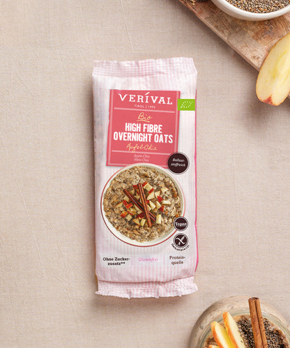 Eine Packung Bio High Fibre Overnight Oats Apfel-Chia der Verival Bio GmbH steht auf einer beigen Fläche, daneben befinden sich Schalen mit Chiasamen und Apfelscheiben, die auf dieses ballaststoffreiche Frühstück ohne Zuckerzusatz hinweisen.
