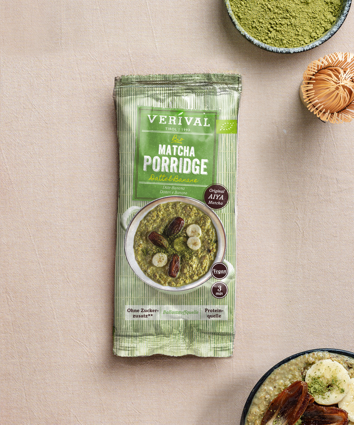 Eine Packung Verival Bio GmbH Bio Matcha Porridge Dattel-Banane steht auf einer beigen Fläche, daneben eine Schale mit grünem Pulver und ein Teil einer veganen Porridge-Schale mit Obst.