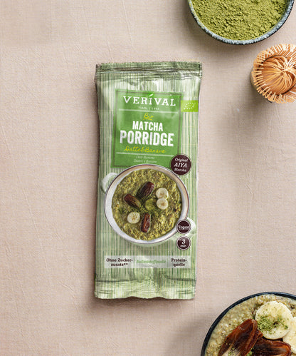Eine Packung Verival Bio GmbH Bio Matcha Porridge Dattel-Banane steht auf einer beigen Fläche, daneben eine Schale mit grünem Pulver und ein Teil einer veganen Porridge-Schale mit Obst.