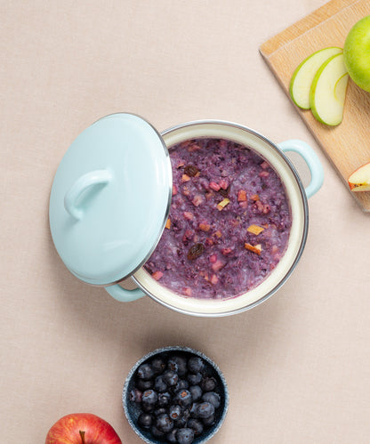 Ein farbenfroher Riess Porridge-Topf mit Deckel ist mit Porridge gefüllt. Daneben liegen auf einem beigen Schneidebrett geschnittene grüne Äpfel, ein ganzer roter Apfel und eine Schale Heidelbeeren.