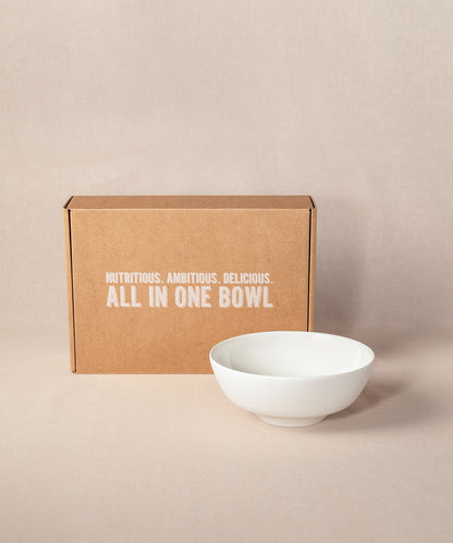 Eine braune Kartonschachtel mit dem Text „Nutritious. Ambitious. Delicious. All in one bowl“ steht aufrecht,  neben einer eleganten, weißen Porridge-Schüssel aus Keramik. Die Schüssel ist Bestandteil des Sets „Breakfast Lover“ von Verival Bio.